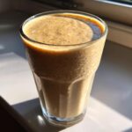 A close-up of a thick, speckled Banana Chip Smoothie served in a tall glass, catching bright sunlight.