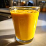 Close-up of a vibrant, thick, orange-yellow glass of the Best Turmeric Smoothie, catching sunlight.