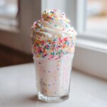 A tall glass filled with a creamy Birthday Cake Shake, rimmed with colorful sprinkles and topped with whipped cream.