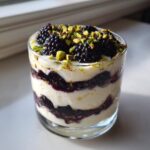 A layered Blackberry Pistachio Cream Delight dessert in a clear glass, topped with fresh blackberries and chopped pistachios.