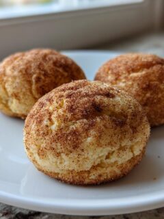 Three freshly baked Brown Butter Snickerdoodles rolled in cinnamon sugar, resting on a white plate near a window.