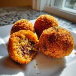 Four golden-brown Cajun Dirty Rice Arancini bites on a white plate, one is broken open showing the rice and filling.