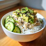 A white bowl filled with California Roll Sushi Bowls ingredients: white rice, shredded crab mix, sliced avocado, and cucumber, topped with sesame seeds.