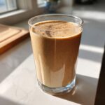 A tall glass filled with a creamy Chai Almond Joy Smoothie, dusted with spice, sitting on a bright countertop.