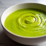 Close-up of bright green Chilled Cucumber Avocado Soup swirled with olive oil in a white bowl.