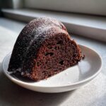 A moist slice of dark Chocolate Bundt Cake dusted with powdered sugar, served on a white plate.