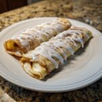Two rolled Cinnamon Roll Protein Crepes drizzled generously with white icing and dusted with powdered sugar.