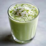 A tall glass filled with a creamy, light green Coconut Key Lime Pie Smoothie, topped with shredded coconut and lime zest.