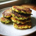 A stack of golden-brown, pan-fried Broccoli Fritters with visible green flecks of broccoli on a white textured plate.