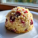 A close-up of a single Easy Cranberry Orange Muffin topped with sugar crumble, sitting on a white plate near a window.