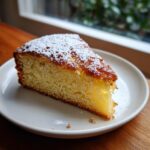 A golden slice of Easy French Butter Cake dusted with powdered sugar on a white plate.