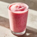 A tall glass filled with a thick, pink Everyday Strawberry Coconut Smoothie with a frothy top.