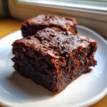 A close-up of two rich, dark Fudgy Sweet Potato Brownies with chocolate chips on a white plate.