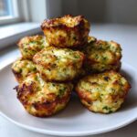 A stack of freshly baked, golden brown Zucchini Tots with visible green herbs on a white plate.