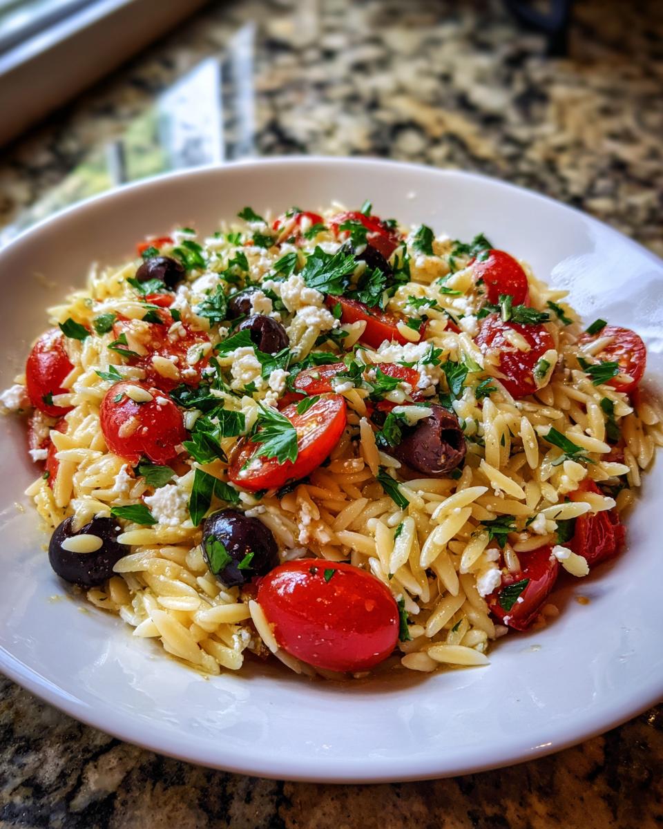 Close-up of a bowl of bright Greek Orzo Salad featuring orzo pasta, halved cherry tomatoes, Kalamata olives, feta cheese, and parsley.
