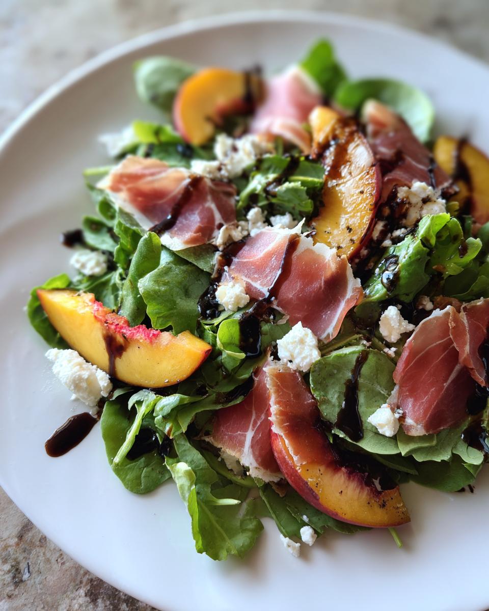 A close-up of a vibrant Grilled Peach Prosciutto Salad featuring greens, grilled peach slices, prosciutto, and feta, drizzled with balsamic glaze.