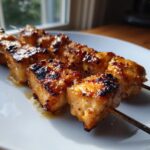 Close-up of perfectly grilled Thai coconut chicken skewers with a sticky, caramelized glaze on a white plate.