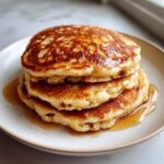 A close-up of a stack of three fluffy Healthy Banana Pancakes drizzled with maple syrup on a light plate.