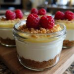 A close-up of a layered High Protein Cheesecake Jar topped with lemon curd, graham cracker crumbs, and fresh raspberries.