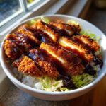 Close-up of sliced, breaded pork cutlet drizzled with dark Tonkatsu sauce over rice and shredded cabbage in a Japanese Katsu Bowl.