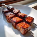 Close-up of two metal skewers loaded with thick, glazed cubes of Korean Beef Skewers, resting on a white plate.