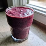 A close-up of a thick, deep purple Lemon Blueberry Avocado Smoothie served in a clear glass by a window.