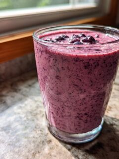 A glass filled with a vibrant purple Lemon Blueberry Avocado Smoothie topped with whole blueberries.