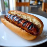 Close-up of a juicy, grilled Marinated Grilled Hot Dog with dark grill marks served in a soft bun on a white plate.