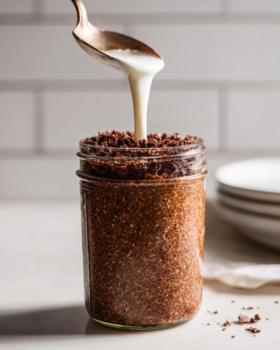 Pouring white cream over chocolate Mocha Overnight Oats topped with chocolate crumbles in a glass jar.