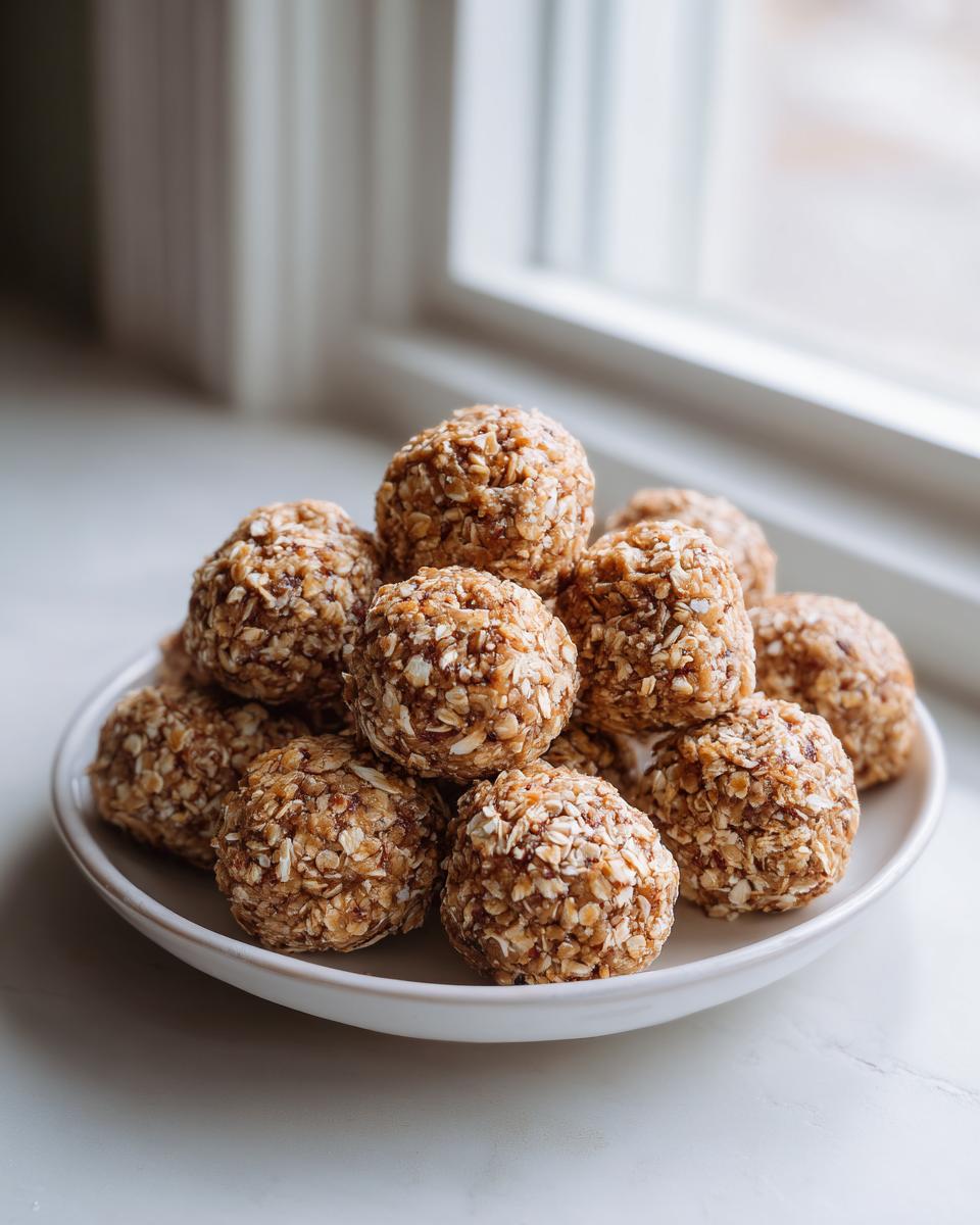 A stack of homemade Peanut Butter Protein Balls coated in rolled oats, resting on a white plate near a bright window.