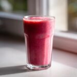 A tall glass filled with a vibrant, thick Pink Glow Smoothie sitting on a sunlit windowsill.