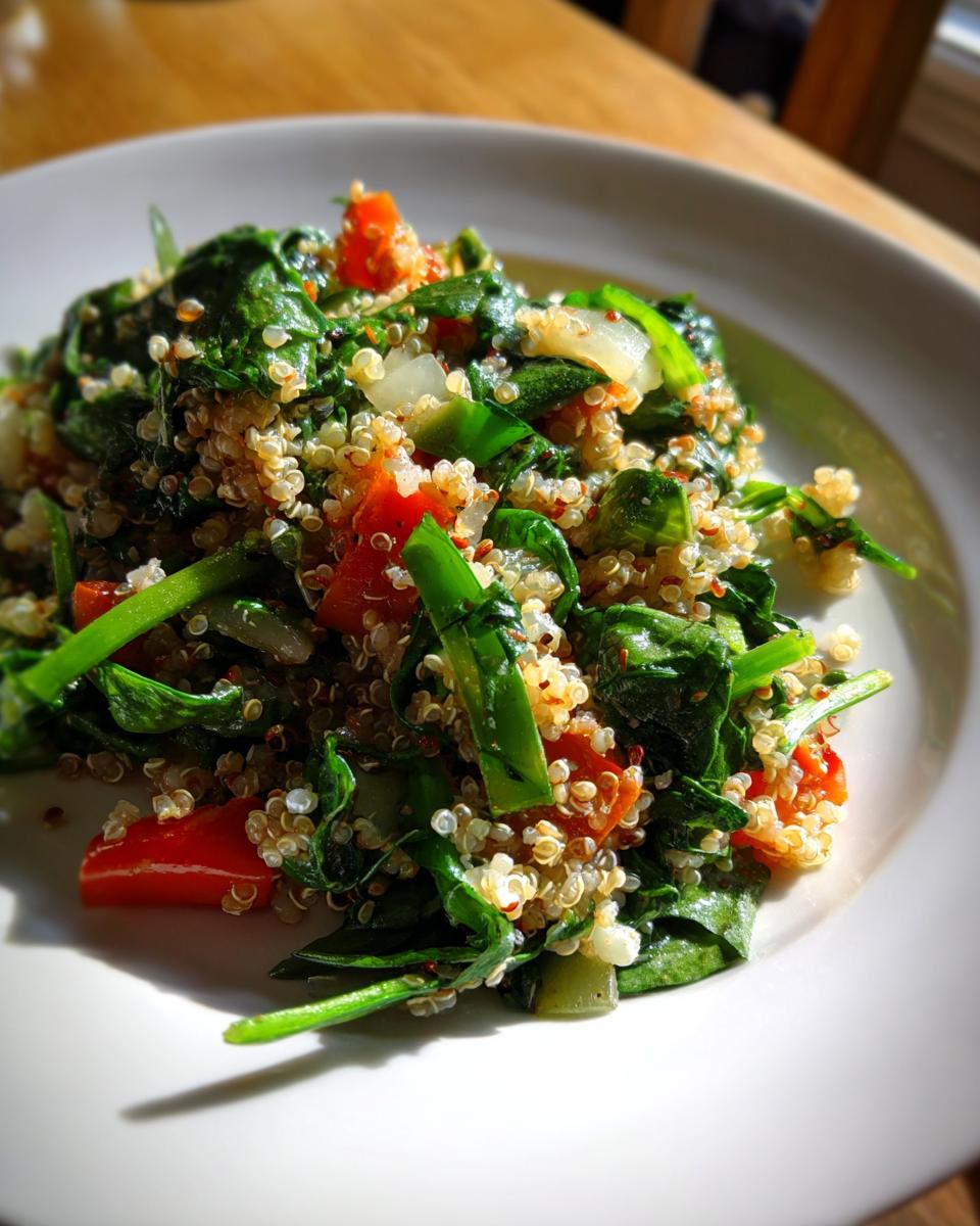 A close-up, sunlit serving of Quinoa Spinach Power Salad featuring quinoa, wilted spinach, diced tomatoes, and onions on a white plate.