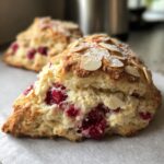 A close-up of a freshly baked Raspberry Almond Scone, studded with bright red raspberries and topped with sliced almonds and powdered sugar.