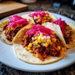 Four Red Chile Chicken Tacos topped with corn and bright pink pickled onions on a white plate.