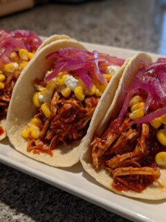Three Red Chile Chicken Tacos topped with corn, cheese, and bright pink pickled onions.