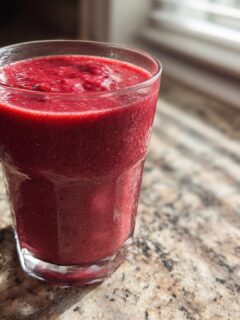 A tall glass filled with a thick, deep red Red Velvet Cake Smoothie sitting on a granite countertop.