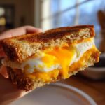 A close-up of a Breakfast Grilled Cheese sandwich with perfectly toasted bread and a runny fried egg yolk dripping over melted cheddar cheese.