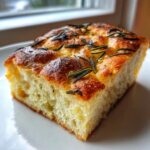 A close-up of a thick, airy slice of Rustic Rosemary Garlic Focaccia with a golden, dimpled crust.