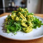 A mound of Smashed Chickpea Avocado Pesto Salad served over fresh mixed greens on a white plate.