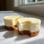 Two smooth Mini Cheesecakes on a marble surface, one with a bite taken out showing the creamy filling and graham cracker crust.