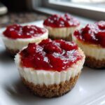 A close-up shot of four individual Strawberry Cheesecake Cups topped with glistening strawberry preserves on a white plate.