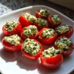A white plate holding several bright red Stuffed Cherry Tomatoes With Herbed Cream Cheese, glistening in the sunlight.