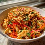 Close-up of a bowl filled with shredded chicken, carrots, and peppers tossed in peanut dressing, topped with chopped peanuts.