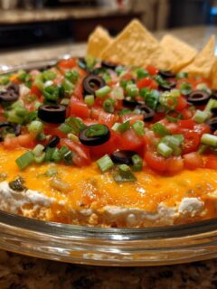 A round glass dish filled with colorful TikTok Boat Dip, topped with melted cheese, tomatoes, olives, and green onions, served with tortilla chips.