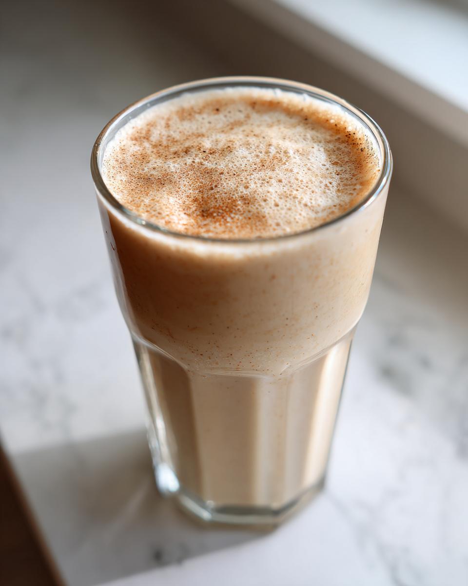 A tall glass filled with a creamy Vanilla Chai Protein Shake, topped with foamy froth and a dusting of cinnamon.