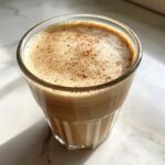 Close-up of a frothy Vanilla Chai Protein Shake in a glass, dusted with spice.