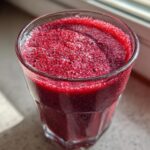 Close-up of a vibrant, deep red Cranberry Smoothie with a frothy top, sitting near a window.