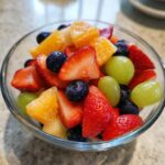 Close-up of a clear glass bowl filled with Fresh Breakfast Fruit Salad including strawberries, blueberries, grapes, and oranges.