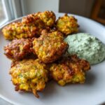 A plate of golden brown Zucchini And Corn Fritters served next to a dollop of green herb sour cream.