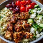 A close-up of a vibrant Greek Chicken Meal Prep Bowl featuring seasoned chicken, quinoa, cucumbers, tomatoes, feta, and olives.